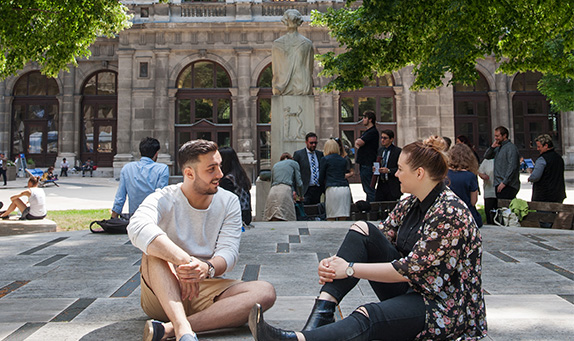 zwei Studenten sitzen im begrünten Arkadenhof der Universität Wien auf dem Steinboden und unterhalten sich.